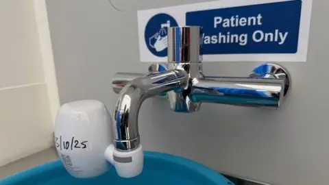 A tap on Wexham Park Hospital's intensive care unit sits above a bright blue plastic bowl. The tap has a white plastic sleeve over it to stop the water from spraying everywhere. The bowl itself sits on a stainless shelf with no drain or pipes in sight.