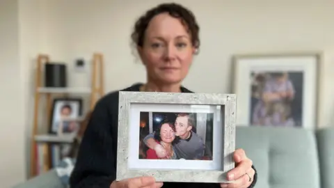 Stuart Woodward/BBC Emma Harley holds up a framed photograph of her and her brother Terry White.  Emma has dark curly hair, she is wearing a dark cardigan, and she is sitting on a green sofa surrounded by other photographs. In the photograph she is holding, she is wearing a red top and grimacing at the camera with her eyes closed. Her brother Terry has an arm around her shoulder and is giving her a kiss on the cheek. He has short shaved hair and is wearing a grey top.