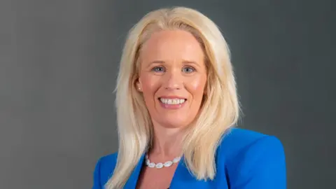 NIAO/PA Dorinnia Carville wearing a blue blazer with black shirt and white necklace. She has blonde hair.