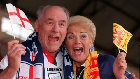 PA Media A man and a woman dressed up in football gear wave flags. They look happy and excited. 