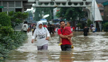people walk through a flooded street in Indonesia, a woman and a man are walking toward the viewer in water that is thigh-high. The man is carrying a toddler.