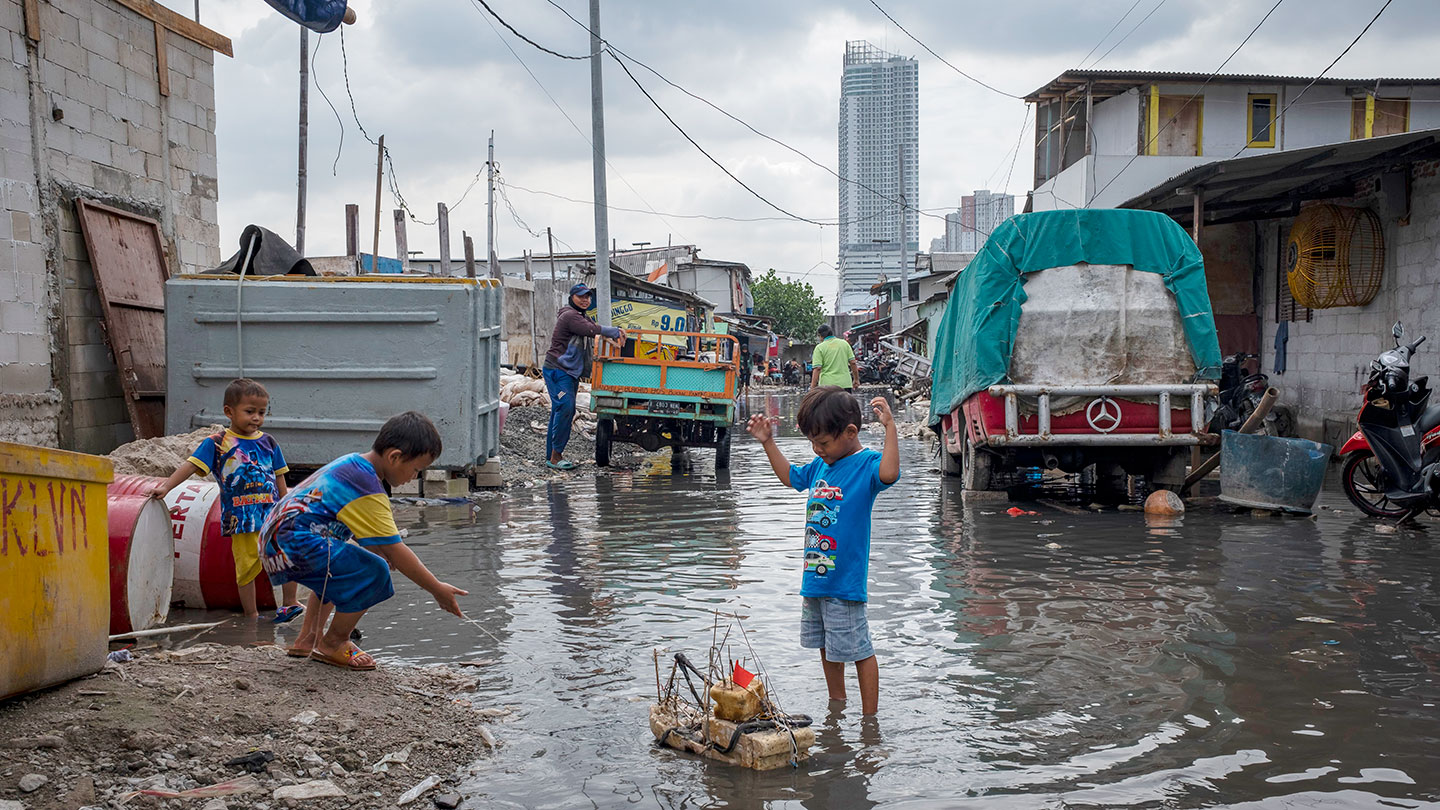 A photo of a boy playing in the middle of a flooded street, he has a small boat assembled from various pieces of plastic, wood and twine. Two other boys stand to the side with him. An adult smiles and watches the kids, standing next to a truck. The water is ankle deep on the young boy and the sky is overcast.