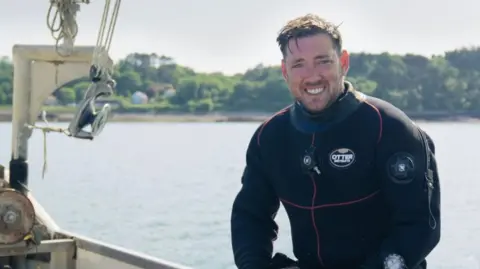 BBC Toby Greatbatch sits on the edge of a fishing boat wearing a wetsuit. He has short brown hair and a beard.