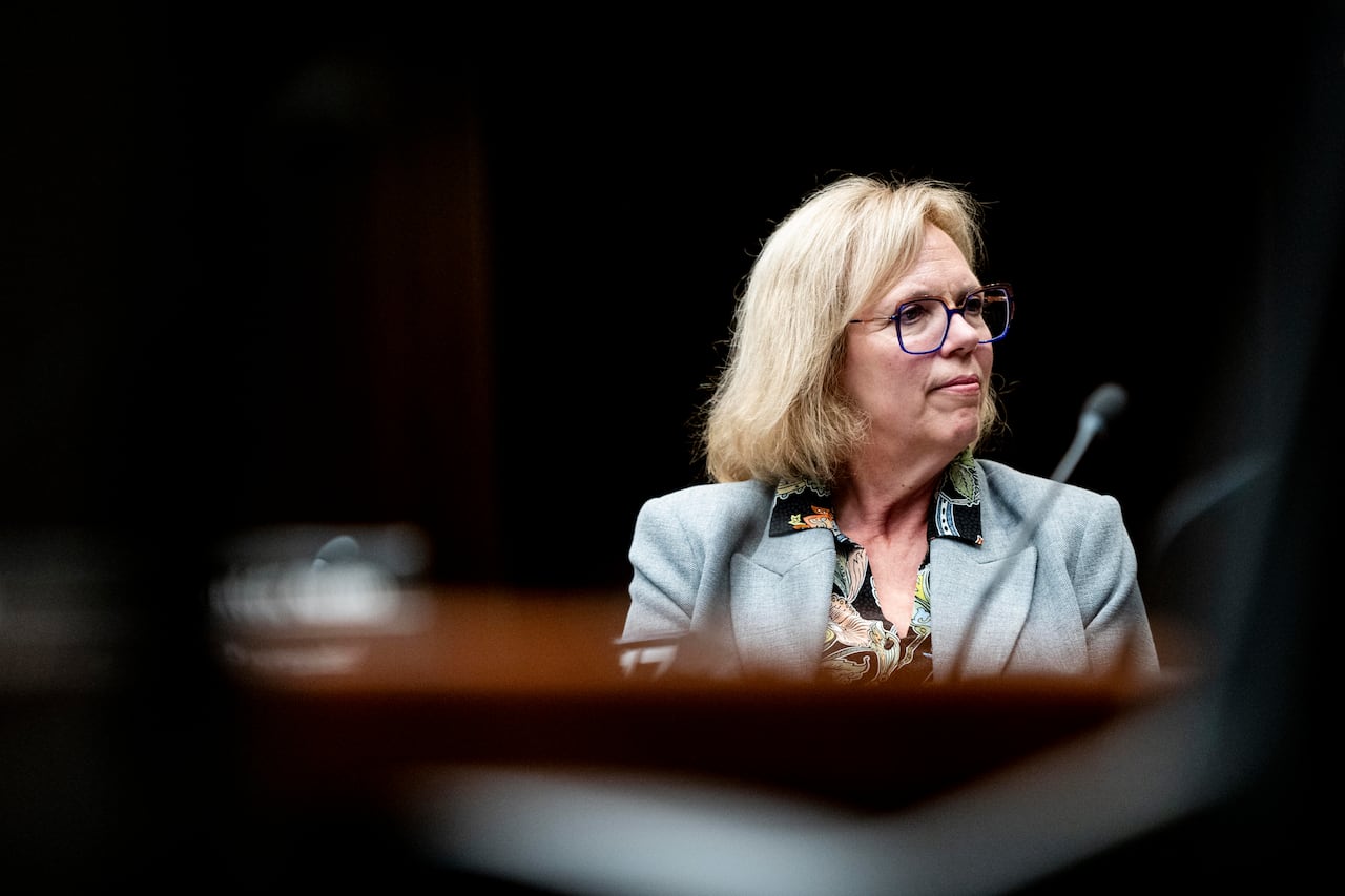 A woman in a grey blazer and glasses sits looking off to the side.