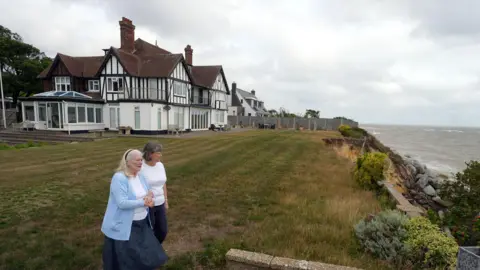 PA Media Jean Flick stands in her garden with her daughter at the edge of a cliff that overlooks the sea on a cloudy day.