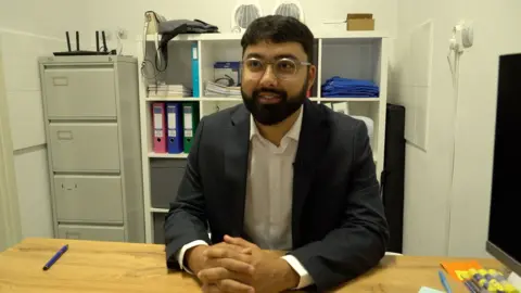Dr Hamza has dark short hair and sits behind a desk. He wearing a black suit blazer and a white shirt.