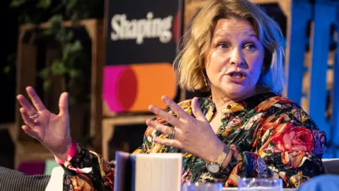 Still Moving Media for Cheltenham Festivals Joanna Page on stage at Cheltenham Festivals, she has short blonde hair and is wearing a multi-coloured floral blouse 