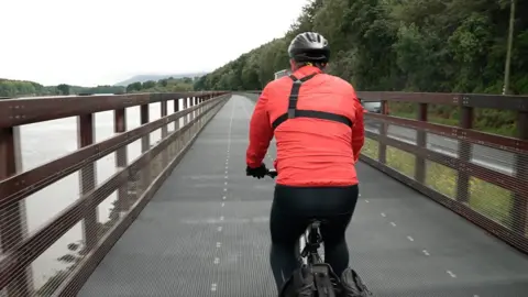 BBC A person from behind cycling along a greenway. A river is to the left and a road to the right. Tall fencing is either side. The person is wearing a black helmet, red rain jacket and black cycling trousers.