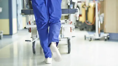 Getty Images Medical professional wearing blue scrubs pushing trolley through a hospital ward