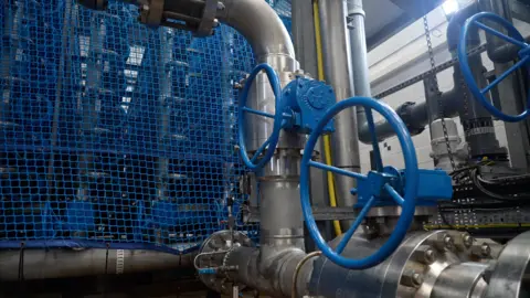 Blue cogs are seen to the right with metal pipes. They are part of the Jersey desalination plant.