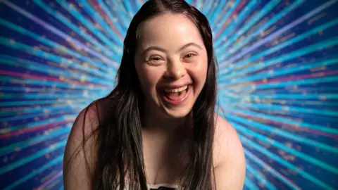 PA Media A woman with brown hair smiling at the camera. There are shining lights behind her. She looks really happy.