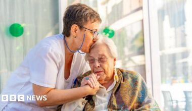 A nurse in a white uniform cares for an elderly woman, who is leaning towards her and has a scarf around her shoulders.