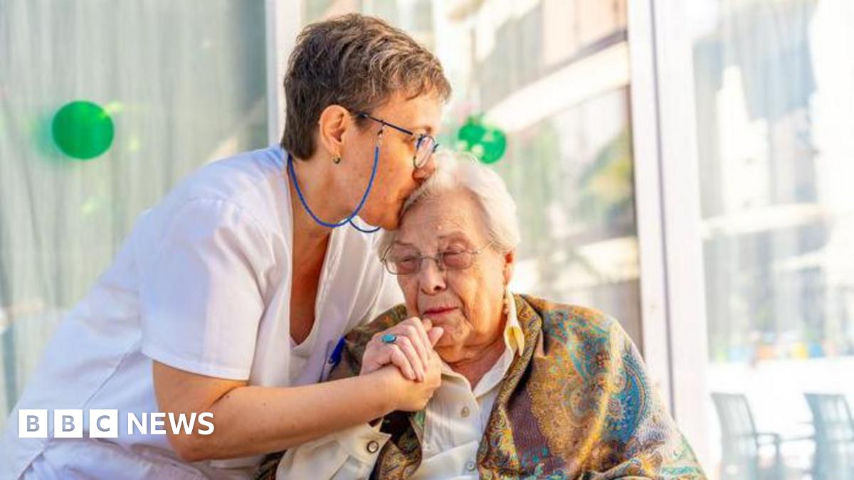 A nurse in a white uniform cares for an elderly woman, who is leaning towards her and has a scarf around her shoulders.