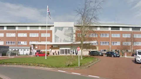 Google Entrance to Cumberland Infirmary. The three-storey, red-brick facade is dominated by rows of windows on each floor, and a large plate-glass entrance in the middle. A Union Flag has been raised on a pole on a small grassy area outside the building; cars are parked in the entrance area.