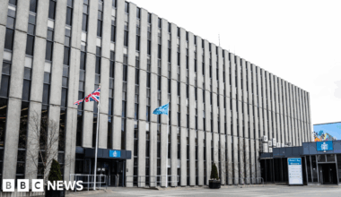 Darlington Town Hall. The large rectangular building is made up of horizontal rows of grey panelling interspersed with windows. Two flags, including a British Union Jack flag, are flying on flagpoles on either side of the entrance.