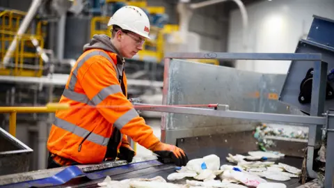 Biffa A man in an orange hi-viz jacket stands at a conveyor belt carrying squashed plastic bottles.