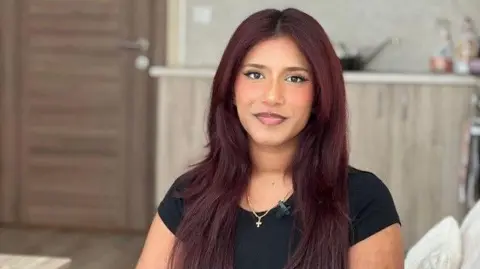 BBC Freya wears a black t-shirt and sit on a cream couch. She has light burgundy-coloured hair. 