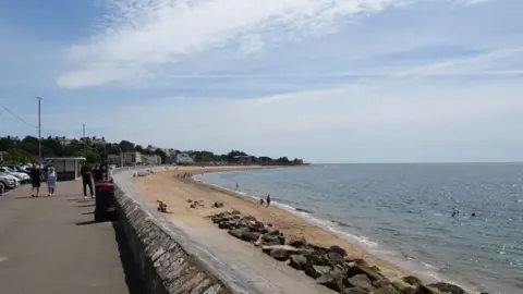 DS Pugh/Geograph The picture shows a seaside promenade running alongside a sandy beach. On the left, there’s a paved walkway with parked cars and a few people walking. A low stone wall separates the walkway from the beach below. The beach itself has golden sand with some scattered rocks near the waterline, and a few people can be seen relaxing on the sand or swimming in the sea. The water is calm, and the horizon stretches out under a bright blue sky with light, wispy clouds. In the background, there are buildings and greenery along the coastline.
