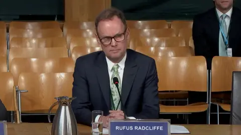 Scottish Parliament Rufus Radcliffe, who has thinning brown hair and black-rimmed glasses, sits with his hands clasped on a wooden table. He is wearing a dark suit, white shirt and green tie. He is sitting behind a placard with his name on it. There are rows of empty wooden seats in the background, with one man in a suit sitting behind him. 