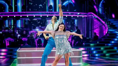 BBC / PA A woman wearing a sparkly blue dress with her arms outstretched. She is stood in front of a man in a light blue top and blue trousers with his arm reaching into the air. They are stood in a ballroom with stairs behind them lit in purple.