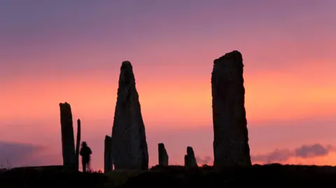 Getty Images A silhouette of an ancient stone circle against a vibrant sunset. The majority of the frame taken up by a gradient sky and the dark shapes of the standing stones and a distant person.