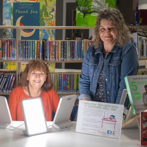 University of Glasgow Two women standing behind a table in a library. The woman on the left is wearing an orange sweater and has reddish-brown hair. She is smiling and looking directly at the camera. In front of her are two white, rectangular lamps with a bright, glowing surface. The woman on the right is wearing a blue denim jacket. She has curly, graying hair and is also smiling at the camera