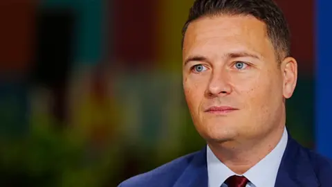 PA Wes Streeting, a man with short dark hair and blue eyes wearing a blue suit, shirt and red tie, looks to the left of the frame