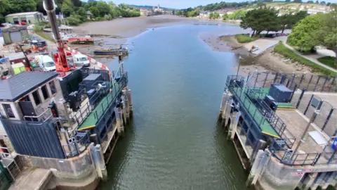 Environment Agency  Truro tide gate, looking up towards the city of Truro. Water is in between the tidal barriers. It is low tide. Trees and green spaces are on land on either side of the water. Properties are in the distance.