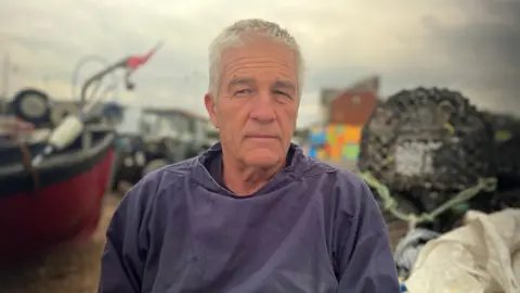 Fiona Irving / BBC A man with short grey hair wearing a blue jumper. He is sat in front of a number of boats which sit on a pebble beach.