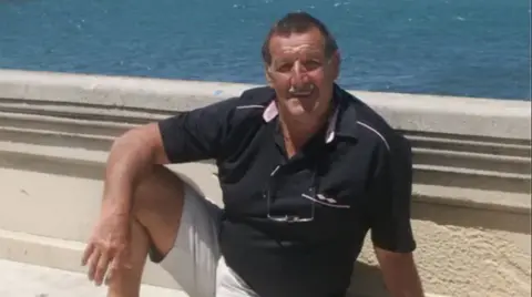 Family photo Colin Flatt wearing a dark polo shirt and white shorts sitting on a concrete bench or wall, with the sea behind him. It is sunny and he is tanned. 