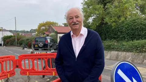 BBC Don Mackenzie stands near Southern Water sewer works in a road. He is bald with white hair at the sides and a moustache and he wears a dark blue cardigan over a pink collared shirt.