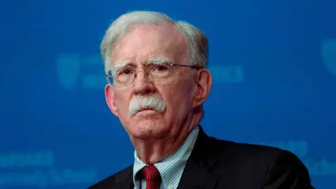 Getty Images A close-up image of John Bolton, who is looking straight ahead. He is wearing glasses, a black blazer, a stripped blue and white shirt and a red tie. 