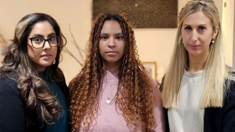 Family handout/PA Wire Three women stand side-by-side with serious expressions on their faces. The woman on the left wears a dark top, has black-rimmed glasses and long brown curled hair; the woman in the centre wears a silver necklace over a pink jumper and has long tightly curled brown hair, and the woman on the right has a white top, and straight blonde hair worn with a plait. 