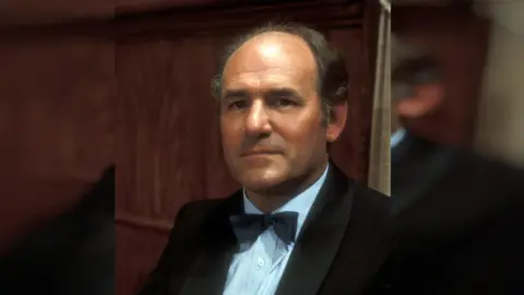 BBC A bald man in a black suit with a bow tie. He is standing behind dark wood. 