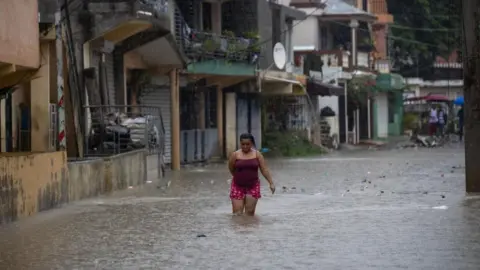 Orlando Barría/EPA/Shutterstock A woman holds up her skirt as she wades through knee-deep water in a flooded street in Santo Domingo. Debris can be seen floating in the water. 