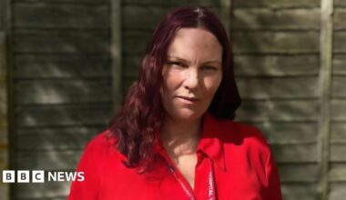 Angie Duffin is wearing a bright red blouse and standing in front of a wooden garden fence on a sunny day. She has long wavy, cherry coloured hair and is wearing a red hospital lanyard round her neck.