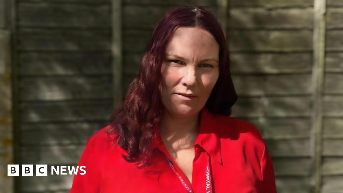 Angie Duffin is wearing a bright red blouse and standing in front of a wooden garden fence on a sunny day. She has long wavy, cherry coloured hair and is wearing a red hospital lanyard round her neck.