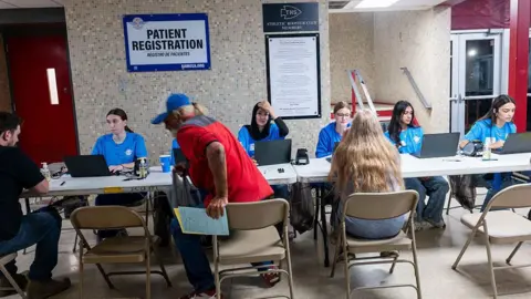 Getty Images Patients register for care at a Remote Area Medical (RAM) mobile dental and medical clinic in Terre Haute, Indiana. Six healthcare workers dressed in blue scrubs sit with laptops across two white tables as three patients sit on foldout chairs
