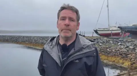A man with light brown hair and stubble, wearing a black waterproof jacket, standing in front of the sea and some boats