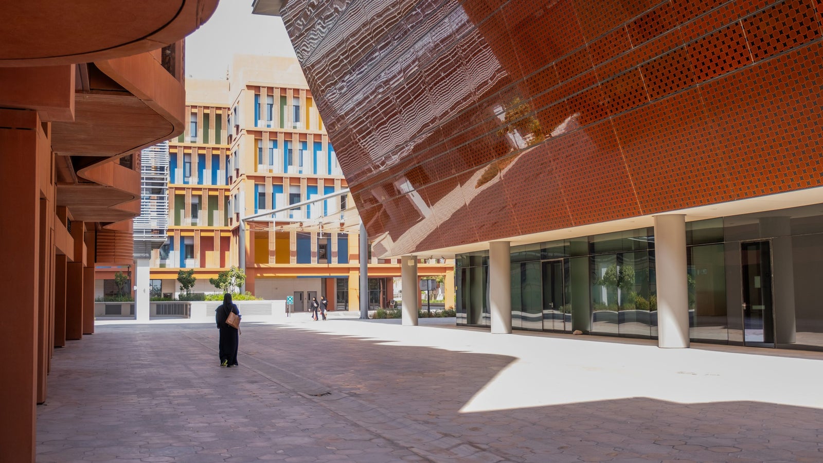 A modern architectural setting with large buildings featuring colorful facades in orange, blue, and green tones. A person wearing a black outfit stands on a paved surface, while another pair of individuals walk in the background under a bright sky.