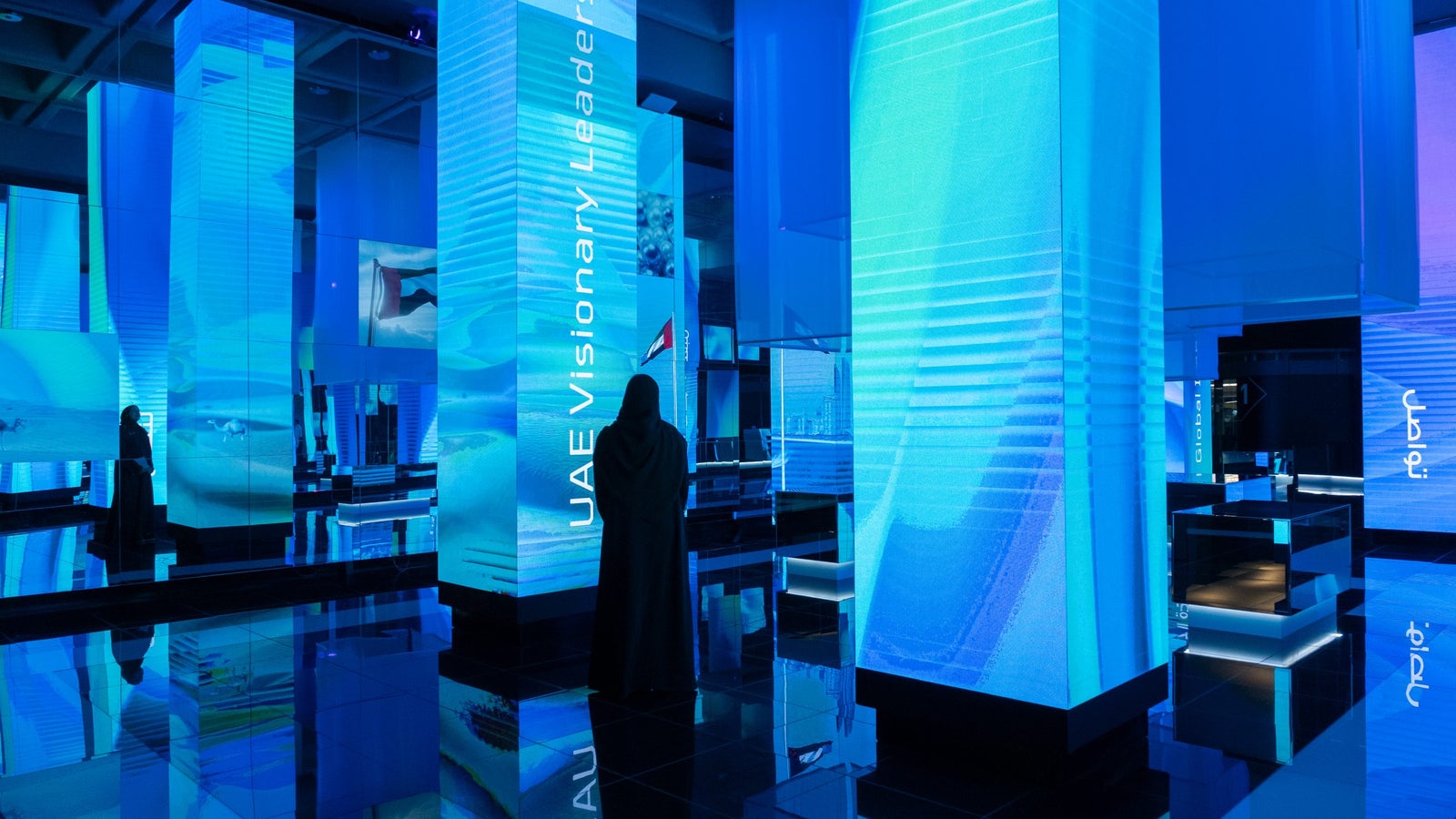 A person in traditional attire stands in a modern, brightly lit exhibition space surrounded by tall, illuminated pillars displaying digital graphics in blue tones. The words 'UAE Visionary Leadership' are visible on one of the pillars, reflecting a futuristic theme.