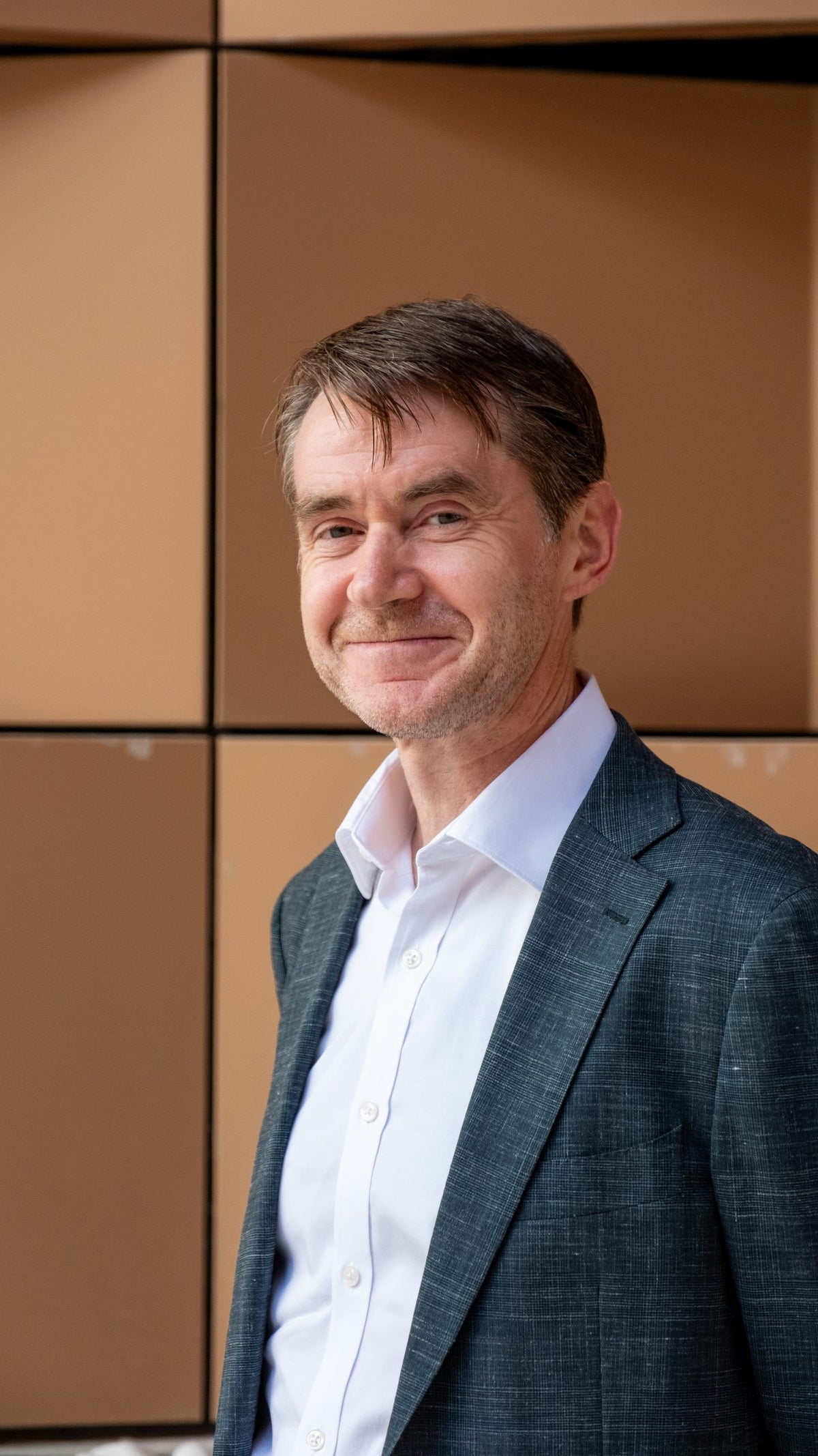 A man with short, neatly styled dark hair smiles gently while wearing a dark patterned blazer over a white shirt, standing against a backdrop of brown geometric panels.