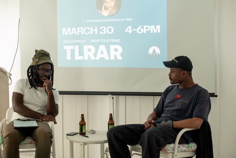two people sit in seats at an artist talk, they both have brown skin adn are wearing hats. 