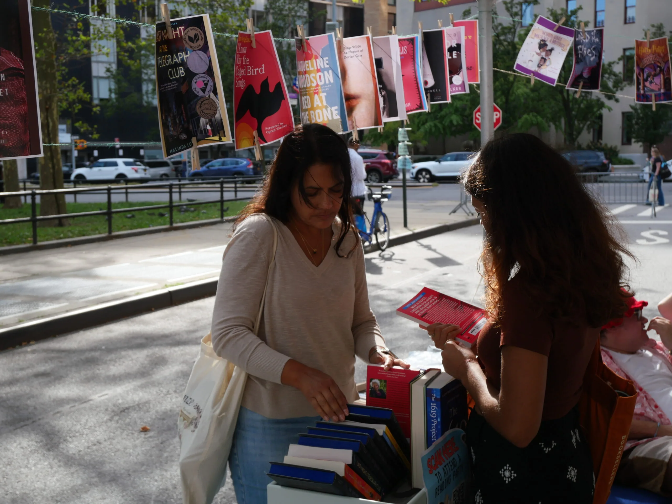 Two women look at books at an outdoor book stall. Behind them, colorful book covers hang on a string above the street while people and a cyclist move through the city background.
