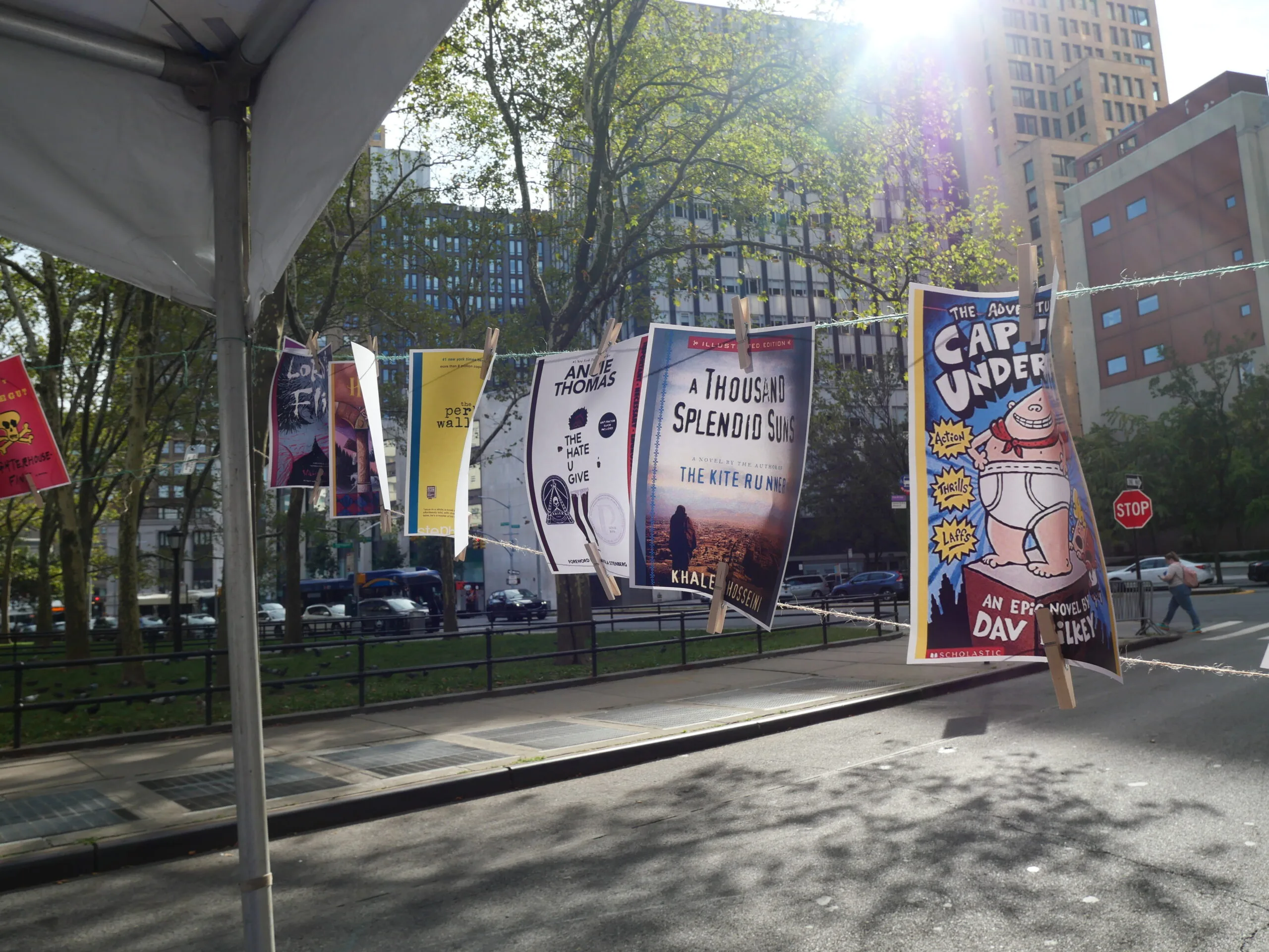 Colorful book covers hang on a line outdoors in a city park, with sunlight streaming through trees and tall buildings in the background. A street and stop sign are visible nearby.
