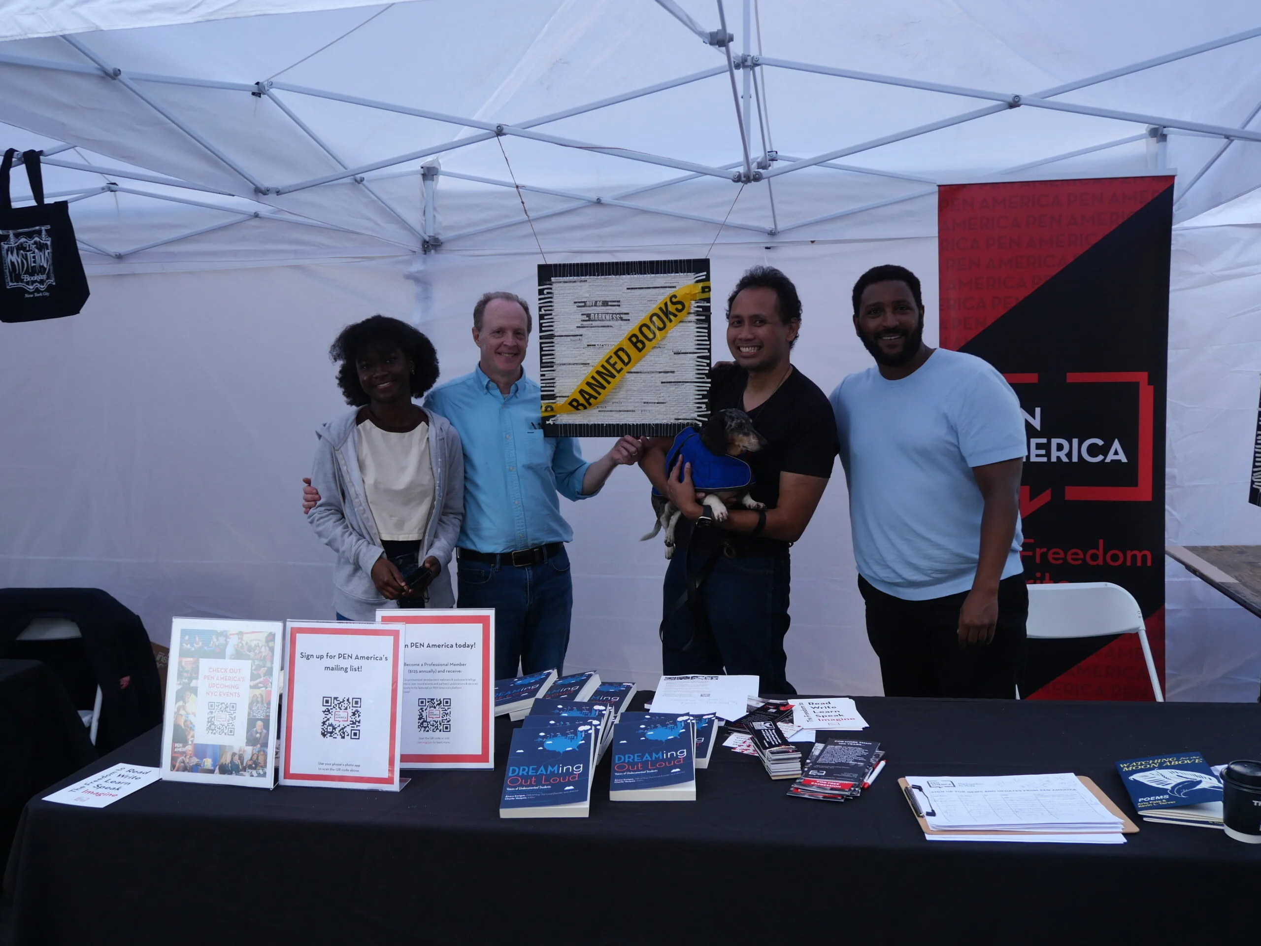 Four people stand and smile behind a table with books, flyers, and signs under a white canopy. One person holds a small dog. A black-and-yellow banner reading Banned Books is displayed behind them.