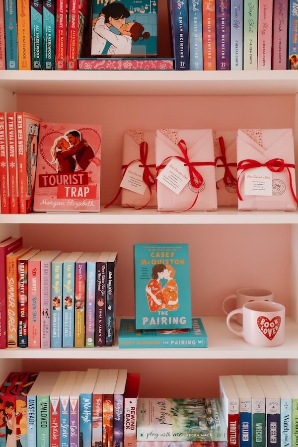 She's Behind Nashville's Sugar and Spice Book Co. - 4 A bookshelf displays colorful romance novels, wrapped books tied with red ribbon, and two white mugs—one featuring "Book Lover" in red and a novel by Christina Duran nestled among the collection.
