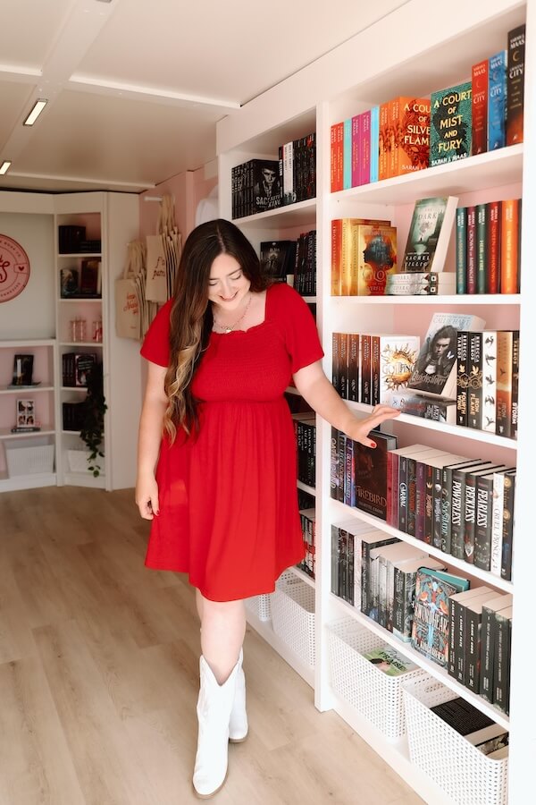 She's Behind Nashville's Sugar and Spice Book Co. - 5 Christina Duran, in a red dress and white boots, stands in a bookstore, gently touching a shelf filled with colorful books.