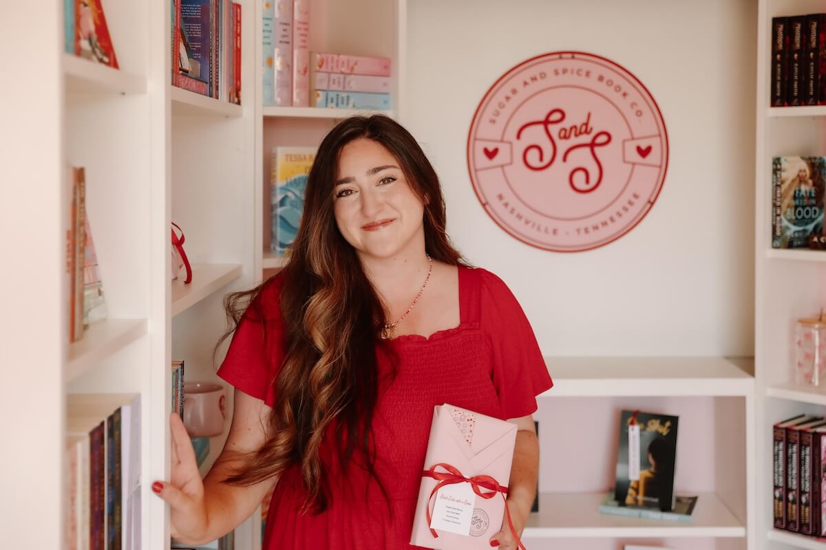 Christina Duran, in a red dress, stands in a bookstore holding a wrapped book. Shelves of books and a wall sign reading “Sugar And Spice Book Co” are visible in the background.