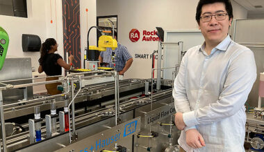 A man wearing a white shirt and standing in a manufacturing environment is smiling as he looks at the camera.